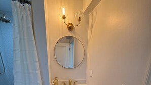 Une salle de bain avec une douche, un lavabo doté d’un robinet doré, un miroir et un luminaire fixé au mur.