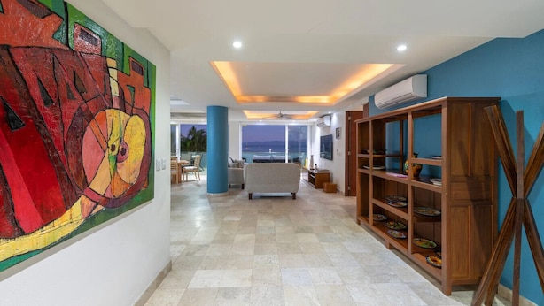 A spacious hallway with a large abstract painting, a wooden shelf with dishes, and a view of the ocean. - Condo Anclote in Corral del Risco (Punta de Mita)
