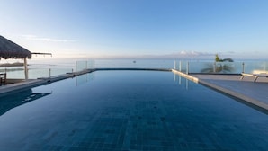 A clear blue infinity pool with a glass barrier, a thatched-roof structure, and a view of the sea and distant mountains. - Condo Anclote in Corral del Risco (Punta de Mita)