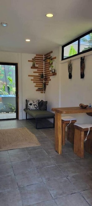 A modern interior with a wooden dining table, a sofa with patterned cushions, a wall-mounted wooden shelf with plants, and a large window. - Mecca- Casita de Reina (Cahuita)