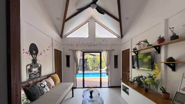 Una habitación amplia con techo alto, ventilador de techo, gran ventana con vista a una piscina, sofá con cojines, una mesita y estanterías con plantas y un televisor.