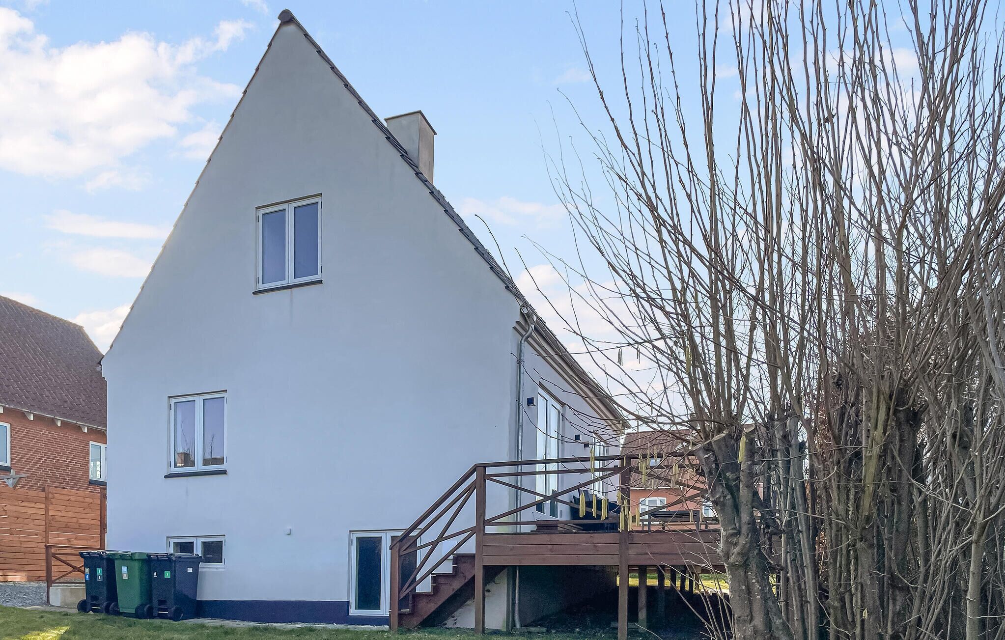 A white building with a triangular roof, a wooden staircase, and a green trash bin.