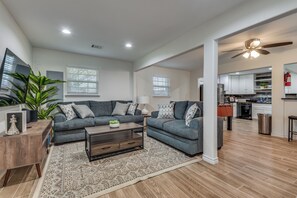 Ein modernes Wohnzimmer mit einer grauen Sofagarnitur, einem Couchtisch und einem Teppich. Im Hintergrund befindet sich eine Küche mit weißen Schränken und einem Deckenventilator.
