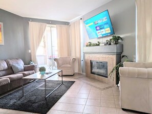 Een woonkamer met een bank, fauteuil, salontafel, open haard en een aan de muur bevestigde televisie.