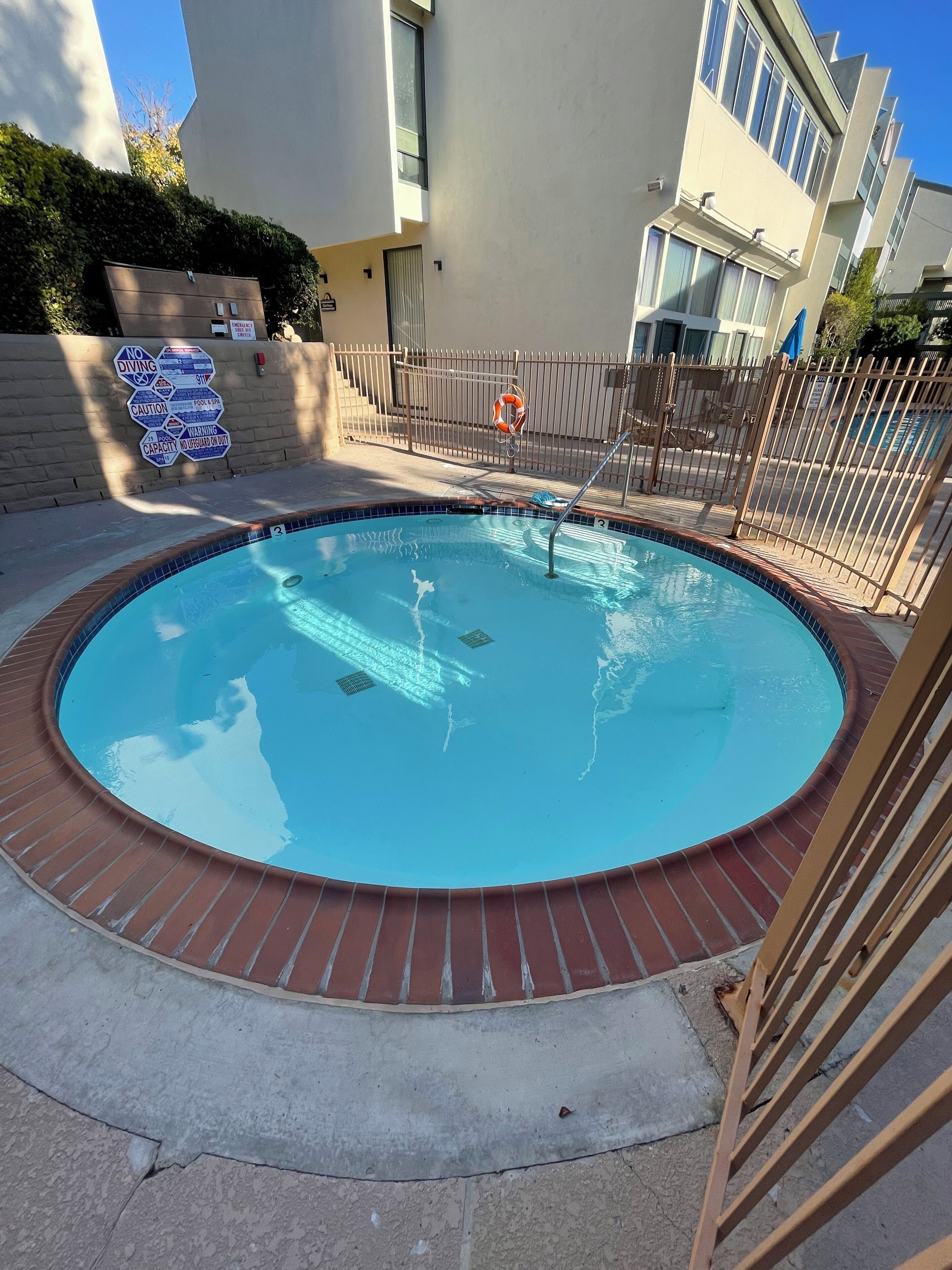 A circular pool with a brick border, surrounded by a metal fence, and a lifebuoy attached to the fence.