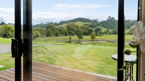Vue depuis une pièce donnant sur une terrasse en bois, avec un champ verdoyant, des arbres et des collines qui se dessinent à l’horizon.