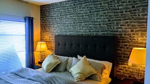 Une chambre avec un mur en briques, une tête de lit capitonnée, deux lampes de chevet et un lit avec une literie à motifs.