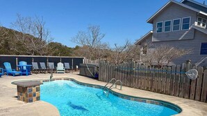 Piscina com deck de madeira, espreguiçadeiras e uma casa ao fundo.