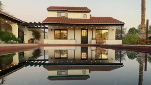 Uma casa de dois andares refletida na água, com uma pérgola e uma palmeira.