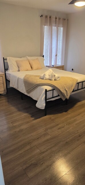 A neatly made bed with a white comforter and beige pillows, a wooden floor, and a window with sheer curtains. - Spacious 4-bedroom house in awesome Davenport with AC (Davenport)