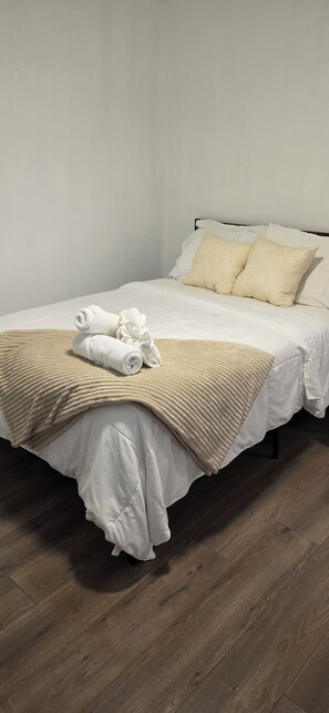 Cama bem arrumada com lençóis brancos e manta bege, acompanhada de toalhas dobradas.