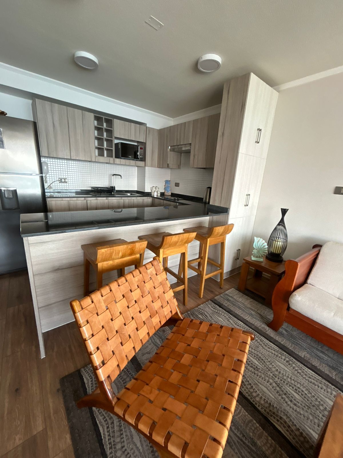 A modern kitchen with a central island, wooden bar stools, and a woven chair.