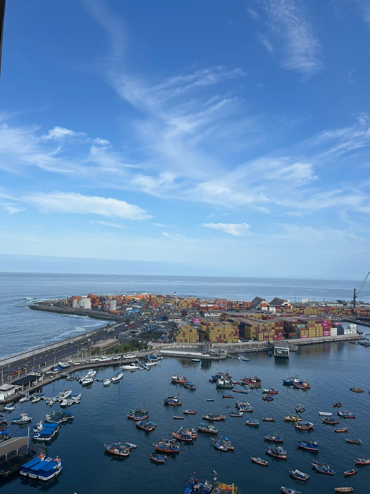 A coastal port with numerous boats docked, a large cargo area with colorful shipping containers, and a clear sky.