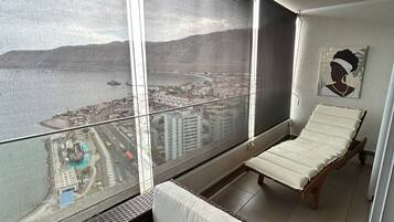 A balcony with a view of a coastal cityscape, a cushioned seating area, and a framed artwork on the wall.