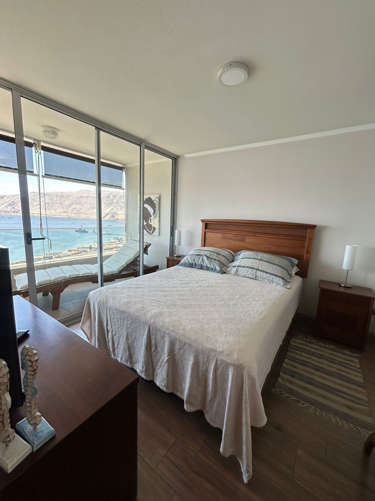 A bedroom with a large bed, a wooden headboard, a nightstand with a lamp, a desk with a keyboard, and a view of the sea through a sliding glass door.