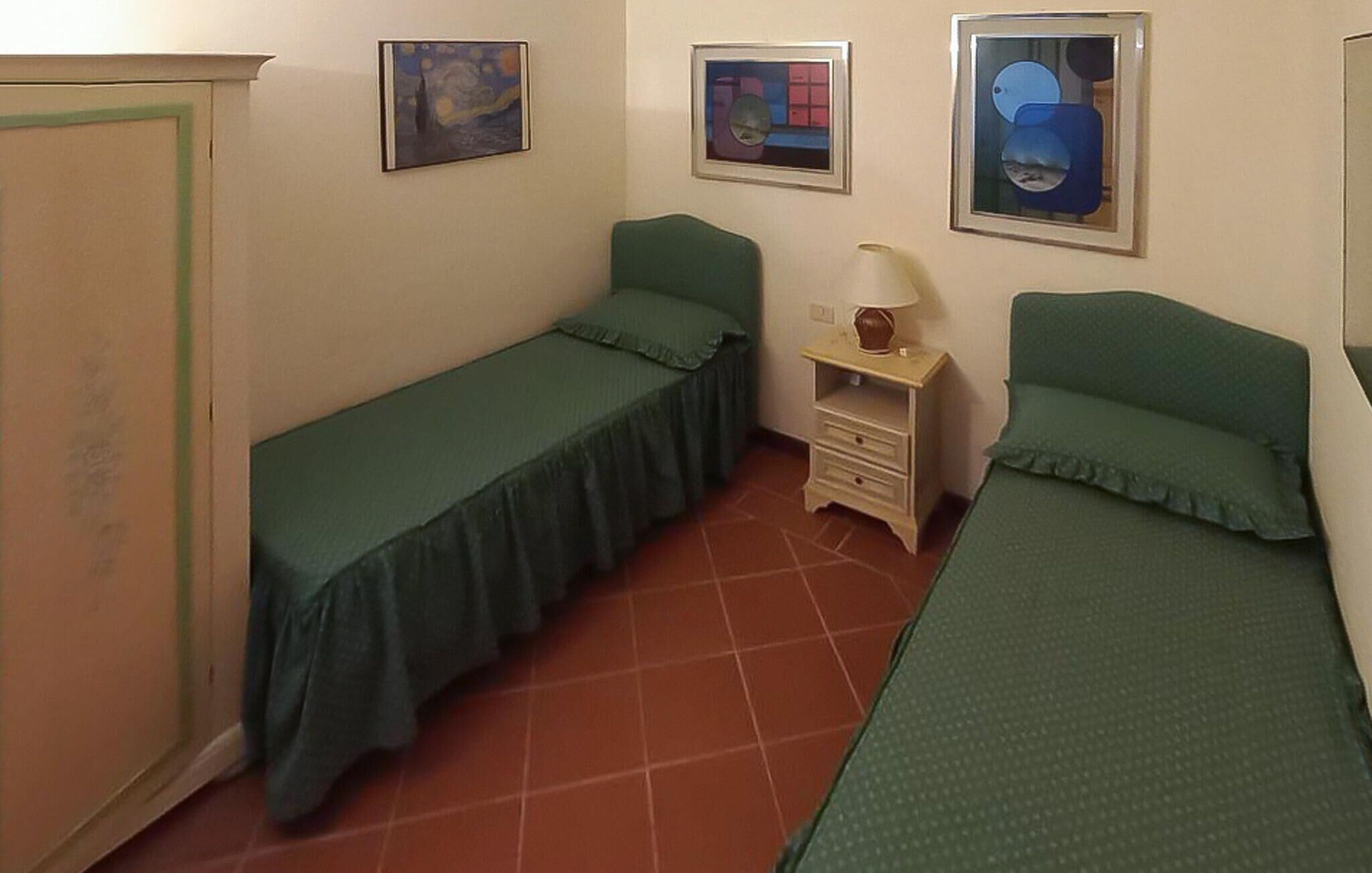 Una habitación pequeña con dos camas individuales, una mesita de noche con lámpara y tres cuadros en la pared.