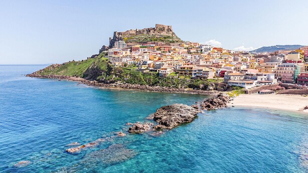 Un pueblo costero con una fortaleza destacada en una colina, aguas azules cristalinas y edificios coloridos.