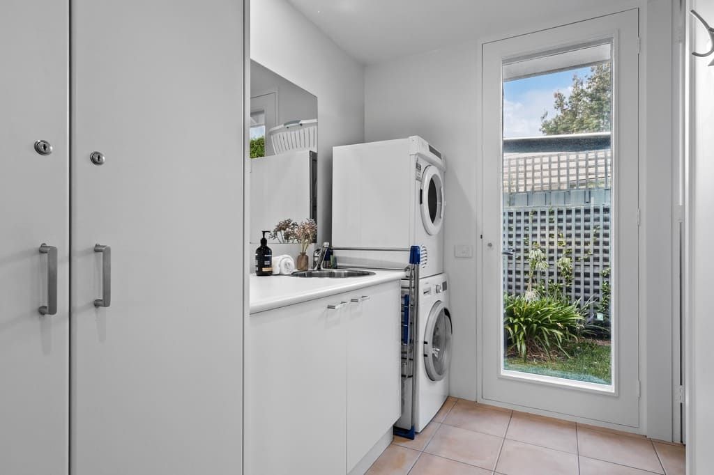 Une petite buanderie comprenant un lave-linge, un évier et une petite fenêtre donnant sur une clôture et de la verdure.