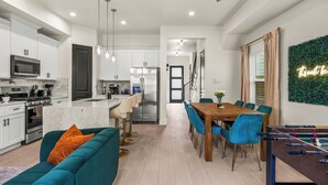 Una cocina moderna con armarios blancos, un microondas y una estufa. Hay una zona de comedor con una mesa de madera y sillas azules. Se ve una mesa de fútbol de mesa al frente.