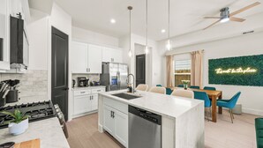 Una cocina moderna con una isla central, electrodomésticos integrados y un comedor con una pared verde y un letrero de neón.