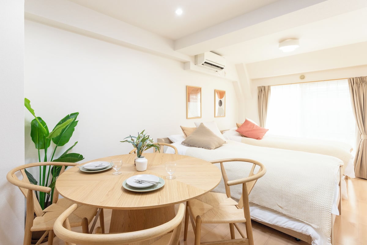 Habitación de hotel con una mesa de comedor redonda de madera, sillas, cama con edredón blanco, ventana grande con cortinas y una planta en maceta.