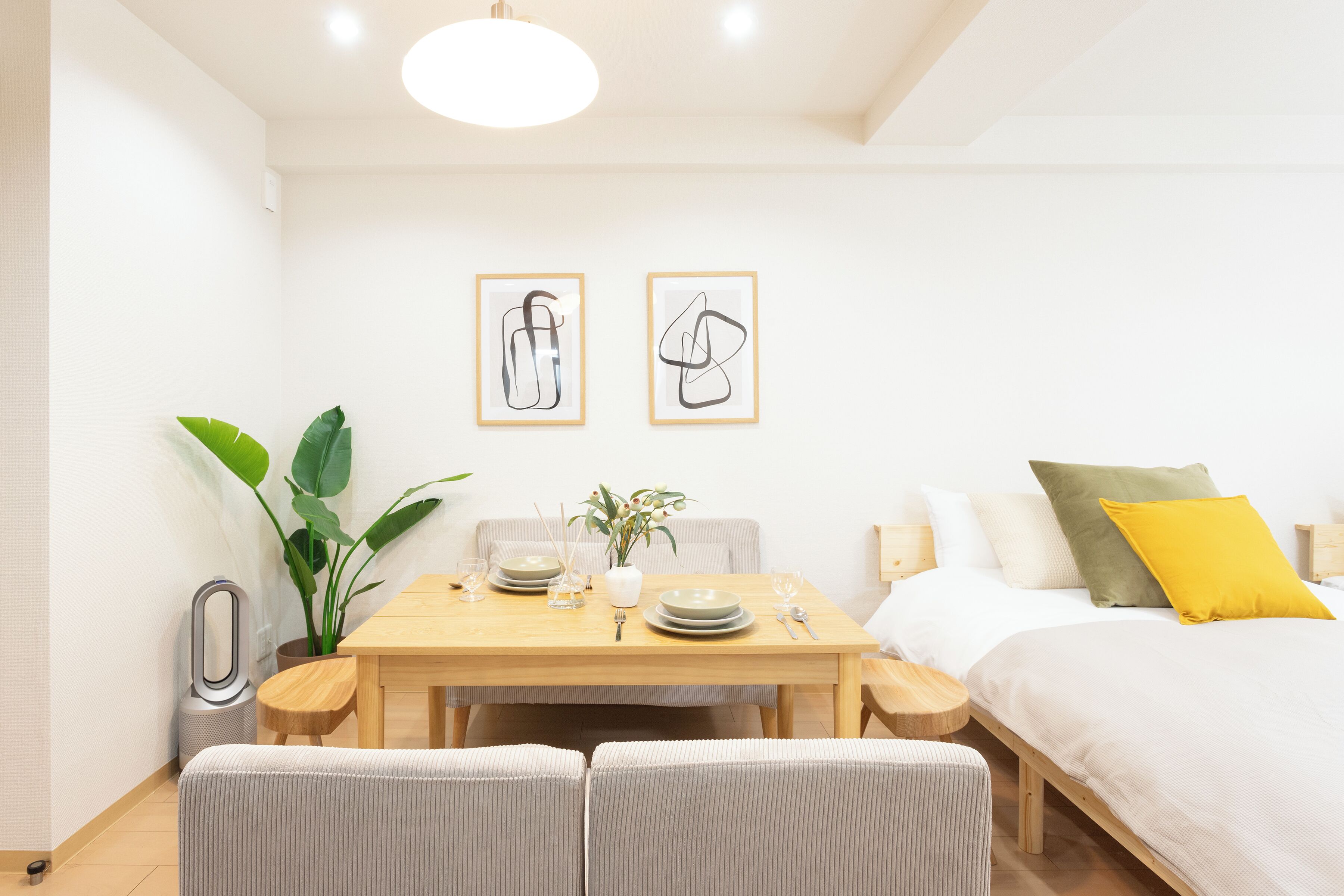 A room with a bed, a dining table with chairs, a potted plant, and two framed artworks on the wall.
