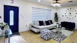 Una sala de estar moderna con sofá, reposapiés y una mesa de centro, una puerta azul y una pared con un reloj.