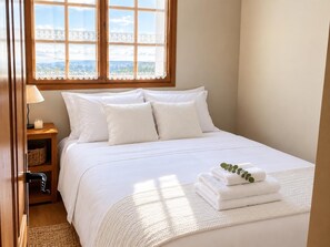 A neatly made bed with white linens, a bedside table with a lamp and a candle, and a window with a view of the landscape. - Duplex ideal for families, swimming pool, fireplace and Wi-Fi (Sainte-Léocadie)