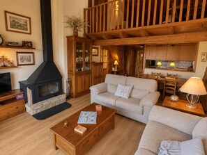 A cozy living room with a fireplace, a sofa set, a wooden coffee table, and a kitchen area in the background. - Duplex ideal for families, swimming pool, fireplace and Wi-Fi (Sainte-Léocadie)