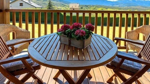A wooden deck with a round table and wicker chairs, a flower arrangement, and a view of mountains. - Duplex ideal for families, swimming pool, fireplace and Wi-Fi (Sainte-Léocadie)