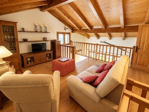 A cozy living room with a sofa, armchair, wooden ceiling beams, a television, and a window overlooking a balcony. - Duplex ideal for families, swimming pool, fireplace and Wi-Fi (Sainte-Léocadie)