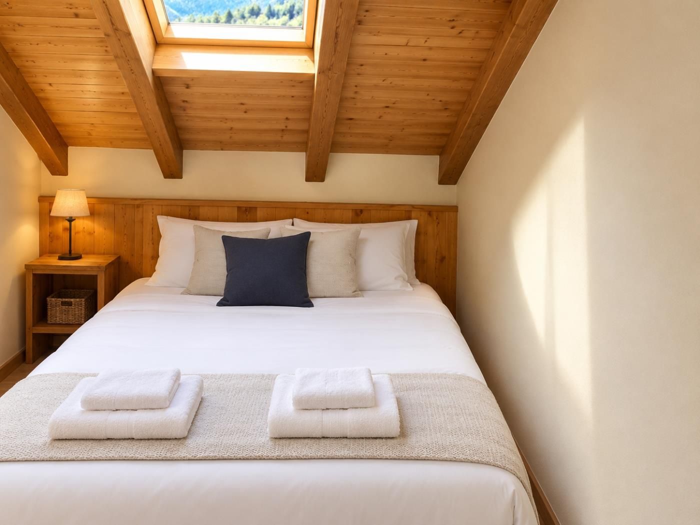 Une chambre avec un plafond en bois, un lit avec une literie blanche, une table de chevet avec une lampe, et une vue sur l’extérieur grâce à une lucarne.