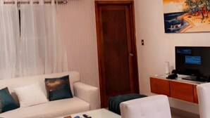 Uma sala de estar com um sofá branco, uma mesa de centro de vidro, uma porta de madeira e um quadro na parede.