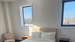Un dormitorio con cama, dos mesitas de noche, una silla y dos ventanas.