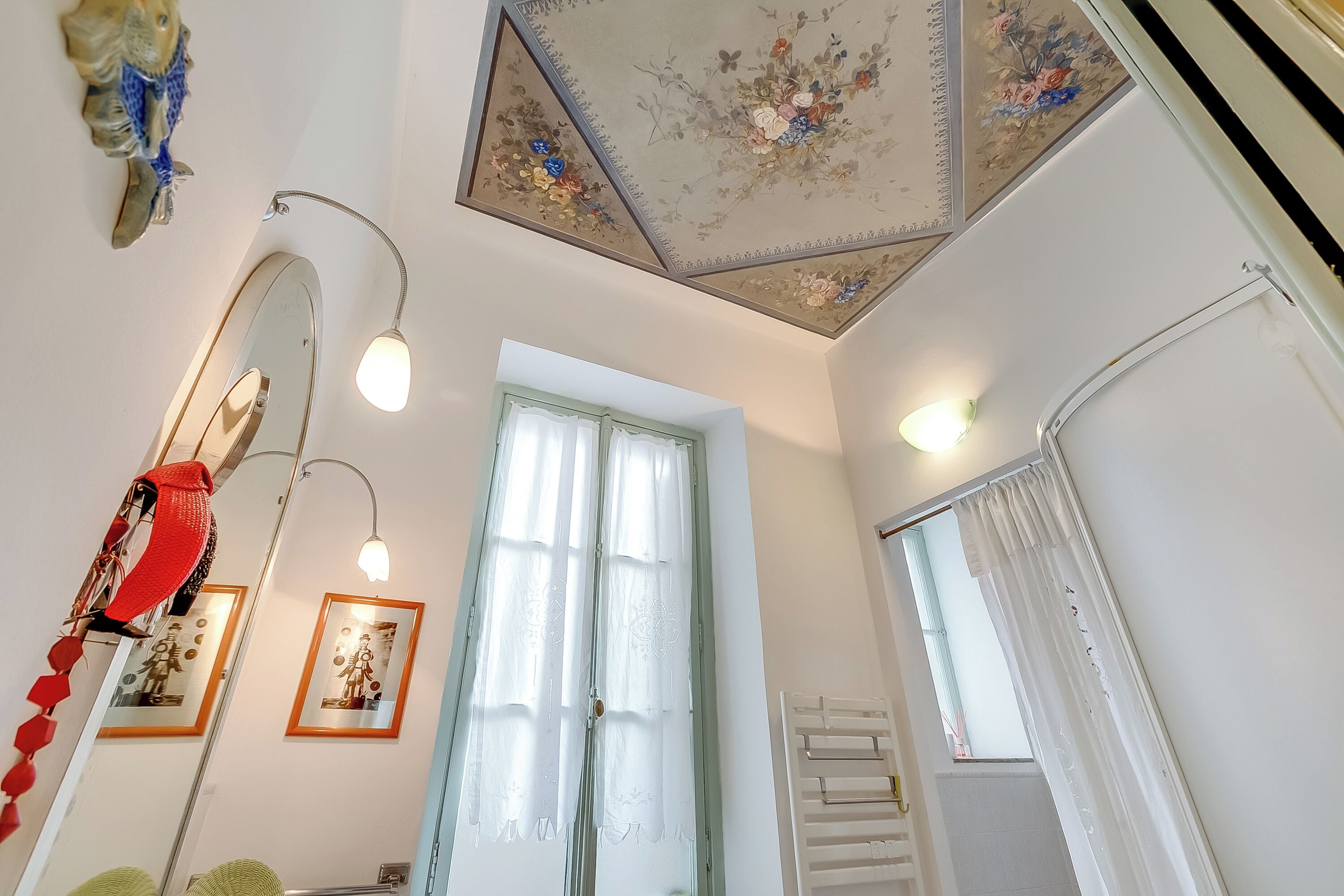 A room with a ceiling with floral patterns, a large mirror, a framed picture, and a window with curtains.