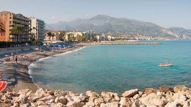Een strand met een rotsachtige voorgrond, een heldere blauwe zee en bergen op de achtergrond.