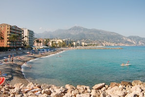 A beach with rocky foreground, a clear blue sea, and mountains in the background. - Clementines 50 Meters From Sea, Roquebrune-Cap-Martin, France (Roquebrune-Cap-Martin)
