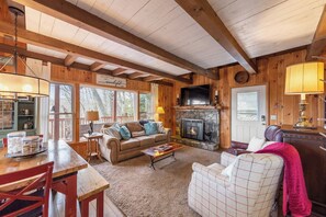A cozy cabin interior with a fireplace, wooden beams, a dining area, and a living room furnished with sofas and a coffee table. - Mountain and River Views - Tucked in the Trees - Wrap Around Porch (Fleetwood)