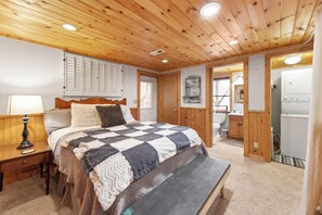 Een slaapkamer met een houten plafond, een bed met een geruit plaid, een nachtkastje met een lamp, en een houten bankje.