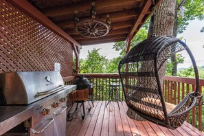 Een houten terras met een barbecue, een rieten stoel en uitzicht op de omgeving.