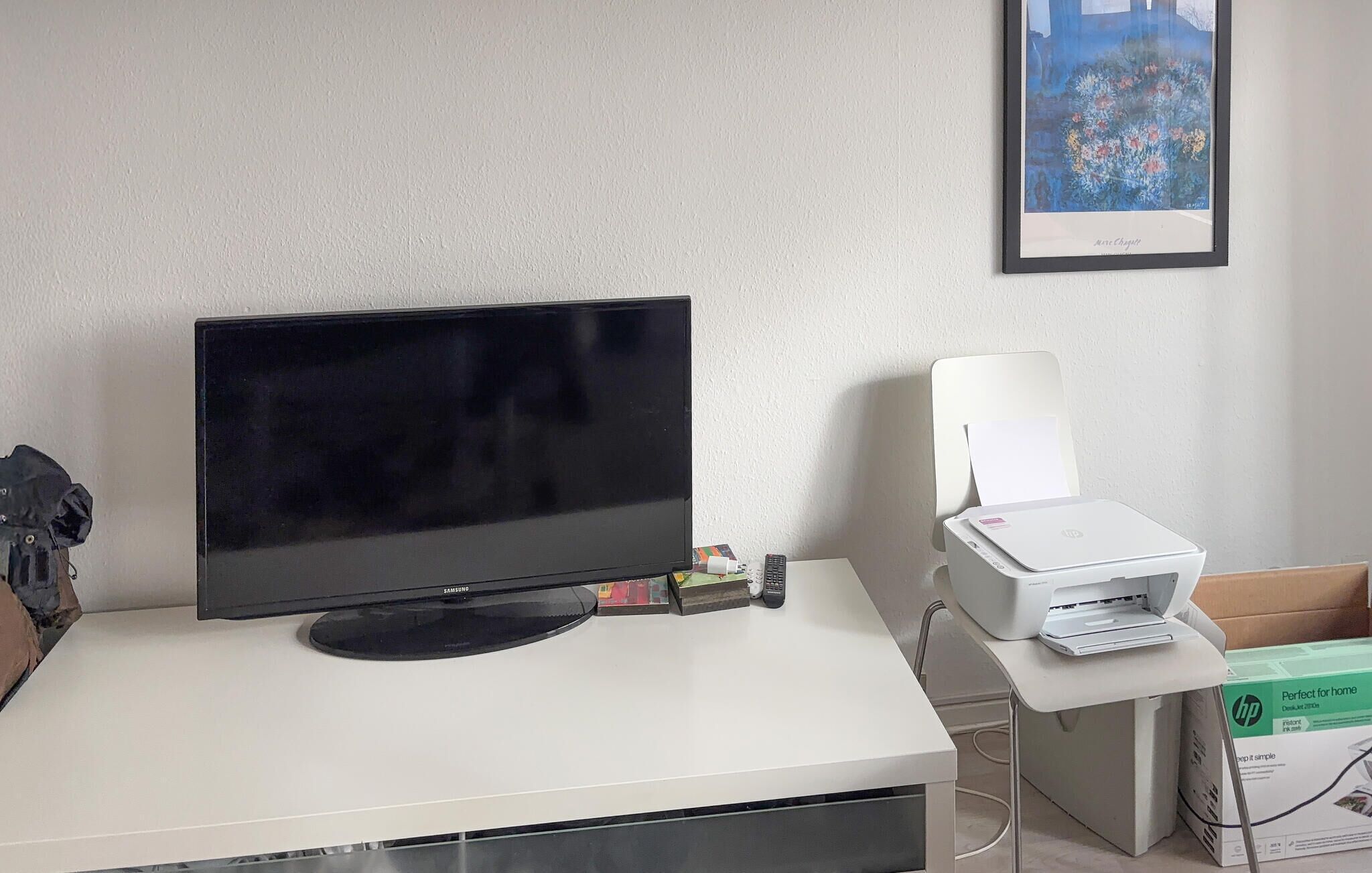 A Samsung monitor on a white desk, a printer, and a framed picture on the wall.