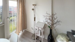 Una stanza con una scrivania bianca, una lampada da terra, un vaso con rami e vista attraverso una finestra con tende.