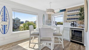Una cocina moderna con armarios blancos, un comedor con una mesa redonda y sillas, y ventanas amplias con vistas al océano y edificios.
