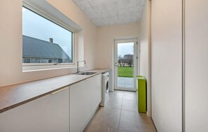 Eine schmale Küche mit Fenster, einer Waschmaschine und einem grünen Mülleimer.