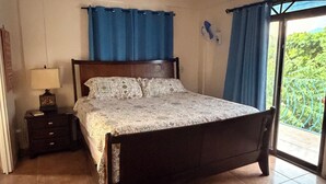 Une chambre à coucher avec un lit, une table de chevet, un ventilateur de plafond et une vue sur la verdure par la fenêtre.