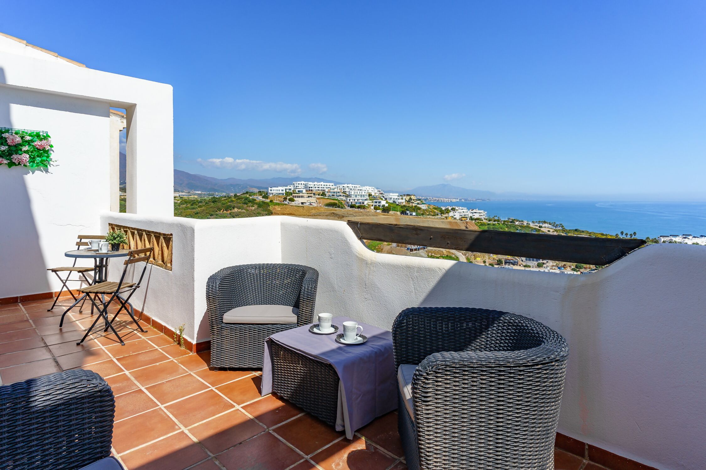 A terrace with wicker furniture, a table with cups, and a view of the sea and buildings.