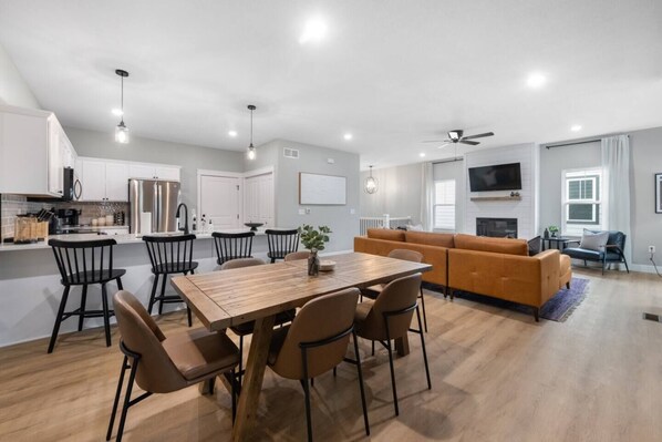 A modern kitchen with white cabinets, a dining area with a wooden table and chairs, a living room with a sofa and TV, and a ceiling fan. - K-State Game House | 3BR w/ Pool + Shuffleboard (Manhattan)