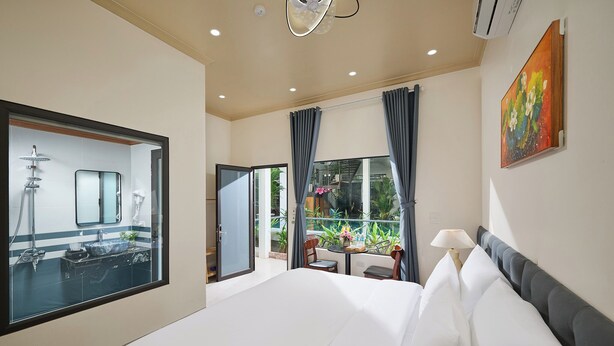 A hotel room with a large bed, a bathroom with a mirror and sink, a balcony with a table and chairs, and a painting on the wall. - Chu Homestay TamCoc (Nam Hoa Lu)