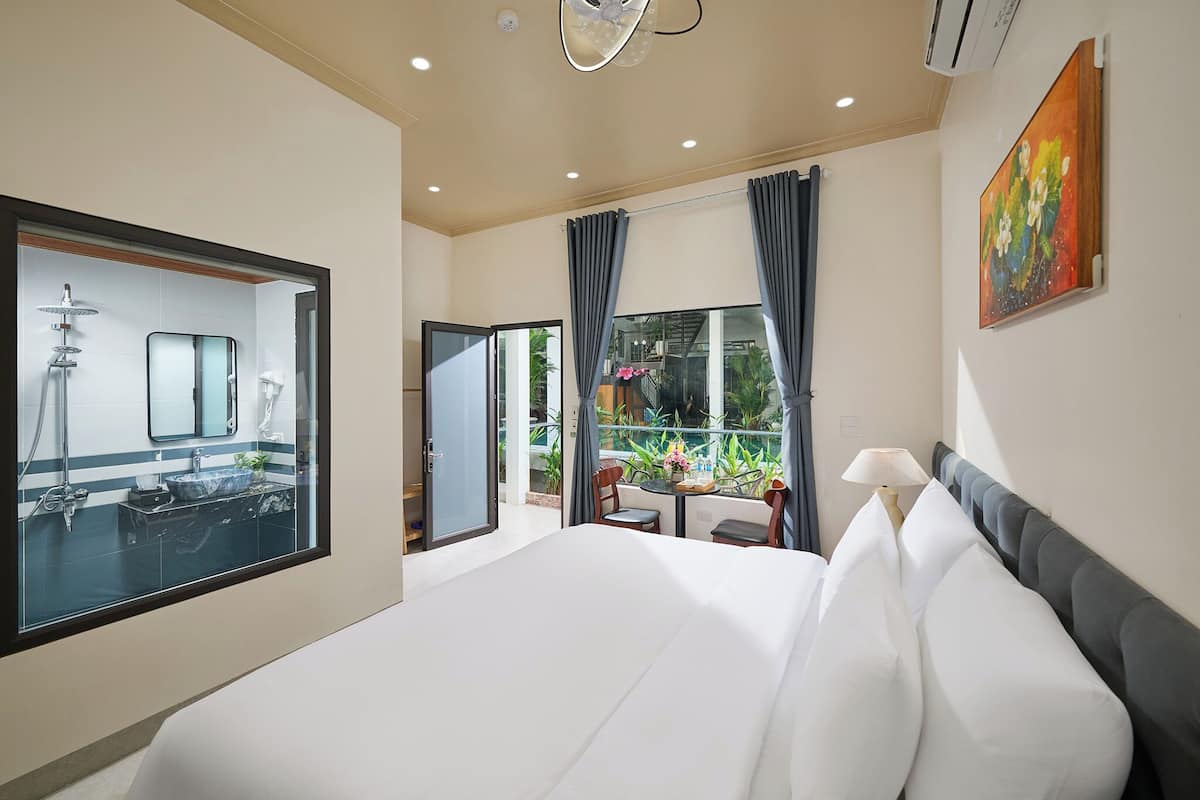 Une chambre d’hôtel avec un grand lit, une salle de bain avec un miroir et un lavabo, un balcon avec une table et des chaises, et un tableau accroché au mur.