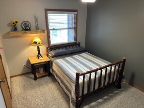 Un dormitorio con cama, una mesita de noche con lámpara, una ventana con persianas y una estantería con adornos.